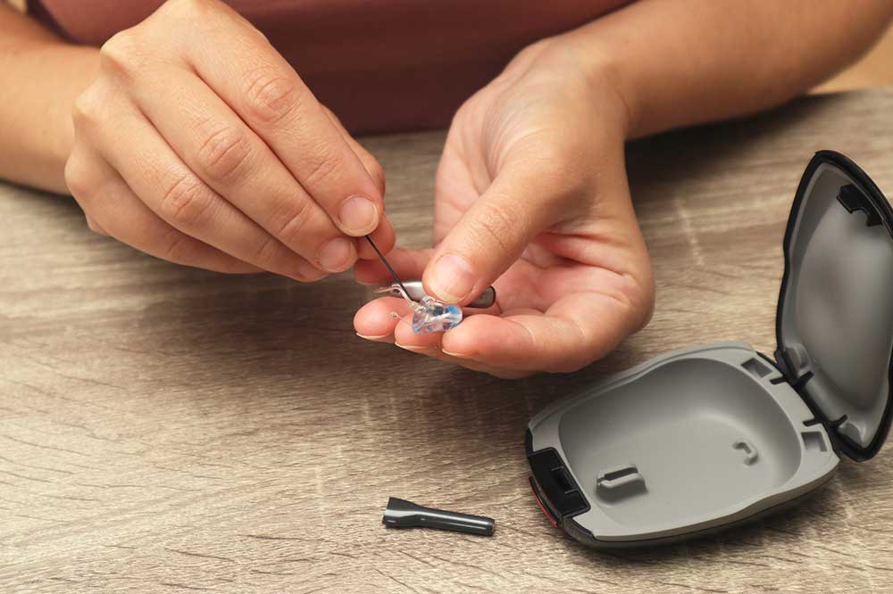 Hands using a wax loop on a hearing aid dome beside an open case, showing hearing aid cleaning tips from Professional Hearing Center in Fremont, NE